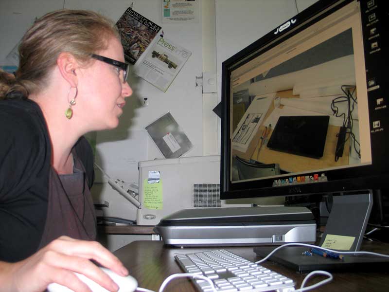 Click the image for a view of: Jessica, at her desk, looking at a picture of Nathaniel's work, next to her work, on his desk, in her studio.